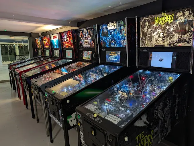 A row of illuminated pinball machines with themed backglasses lines an arcade room, with vintage film posters visible on the walls behind.