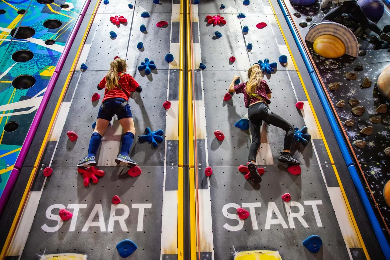 Two children climbing adjacent indoor climbing walls with coloured hand and foot holds.