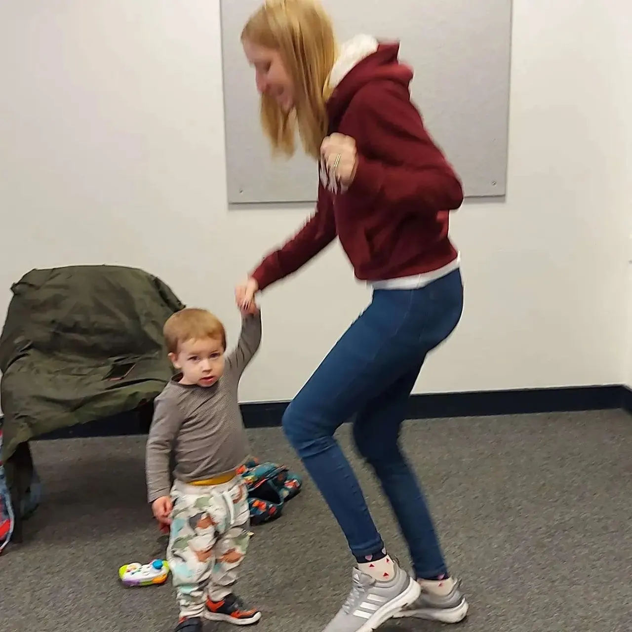 Woman in maroon hoodie dancing and playing with a toddler indoors.