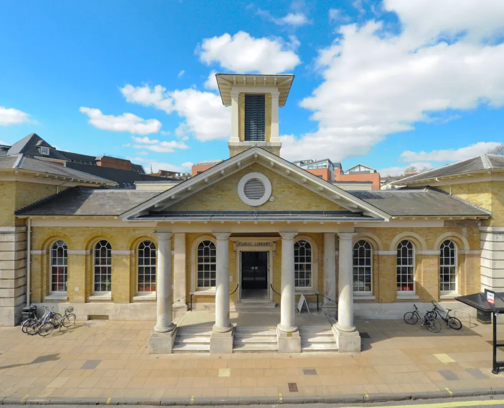 Classical yellow-brick public library building with columned portico, cupola, and bicycles parked outside on a sunny day.