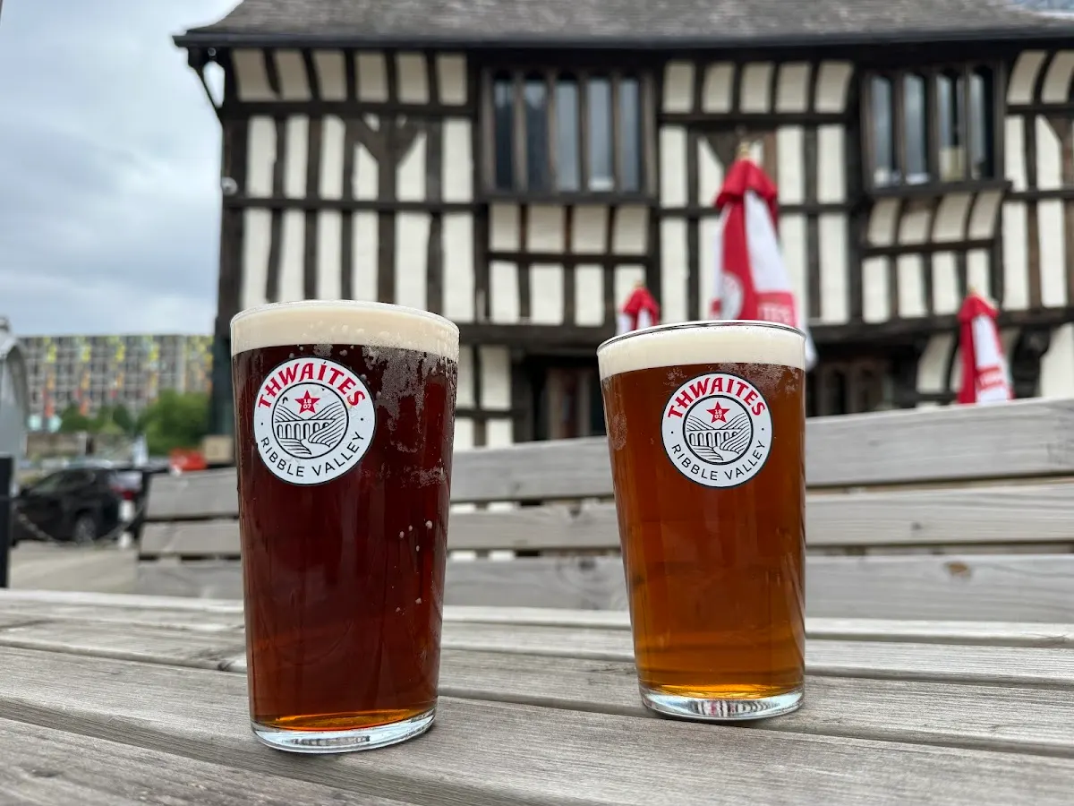 Two pints of Thwaites beer on a wooden table outside a timber-framed historic building.
