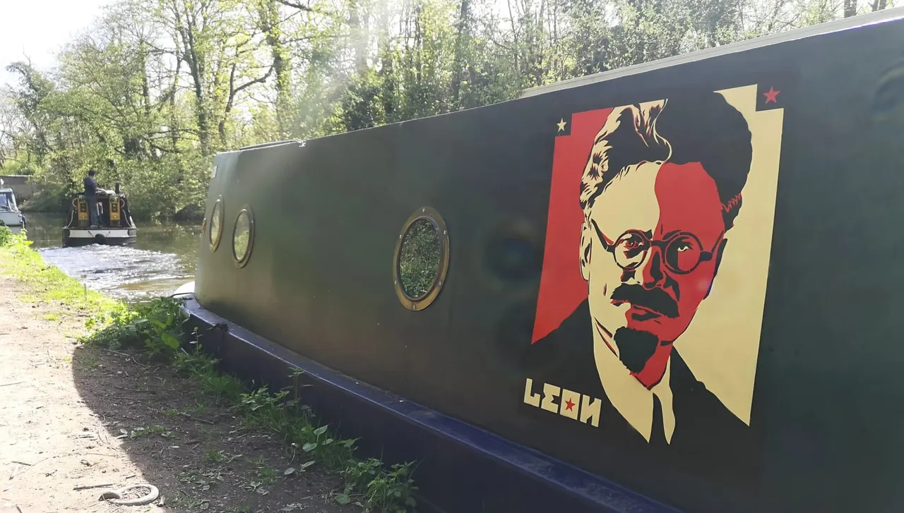 A narrowboat moored on a canal with a bold portrait mural painted on its side, trees reflected in the water behind.