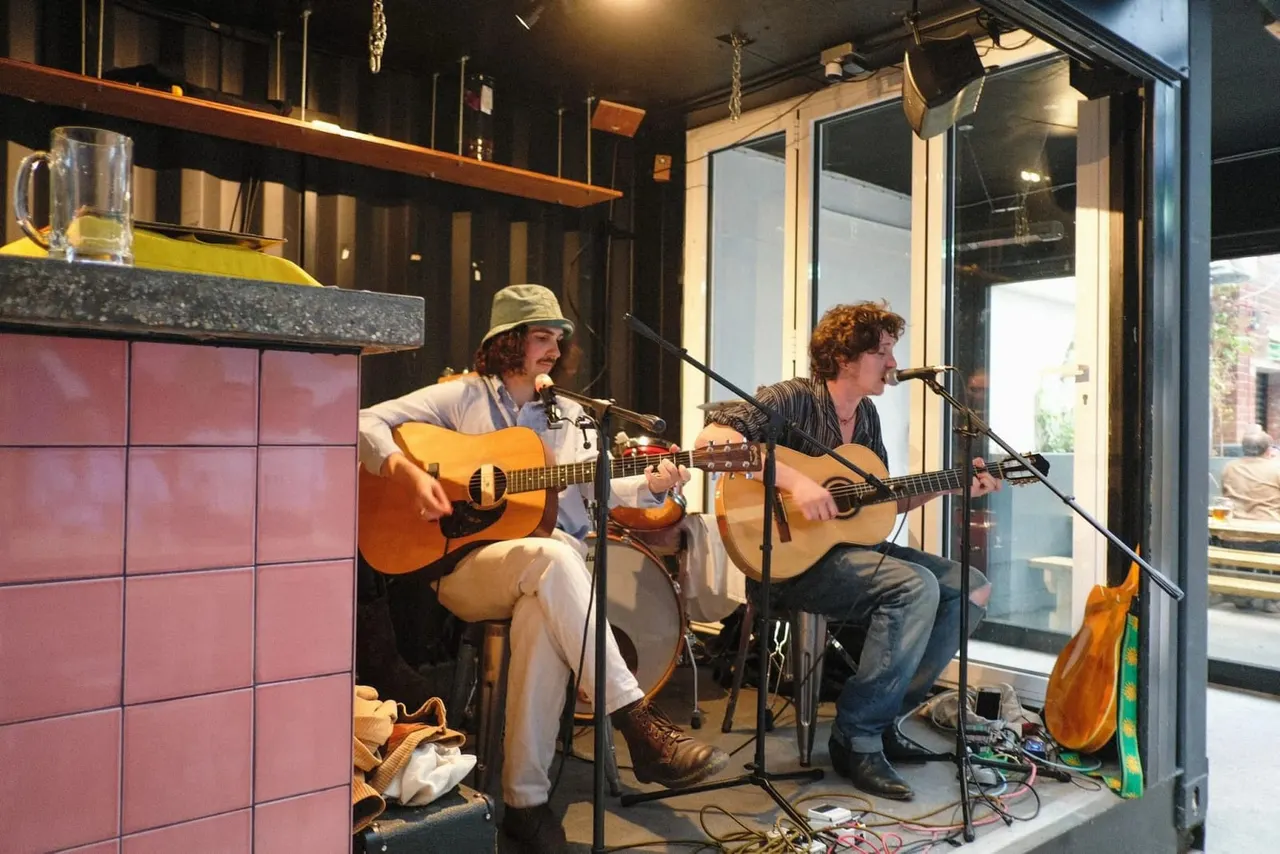 Two musicians with guitars performing at microphones in an indoor venue with modern dark décor and pink tiled walls.