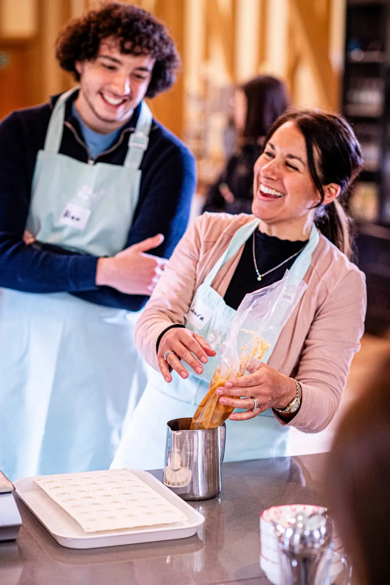 Two smiling people wearing aprons participate in a cooking class, with one pouring batter into a metal jug.