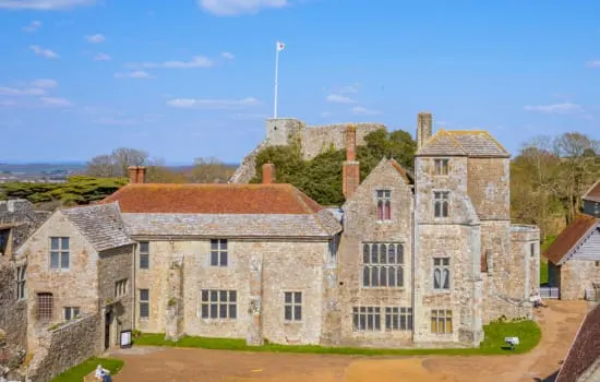 Medieval stone castle buildings with flag flying from fortified tower on hilltop behind, surrounded by green lawns and countryside.