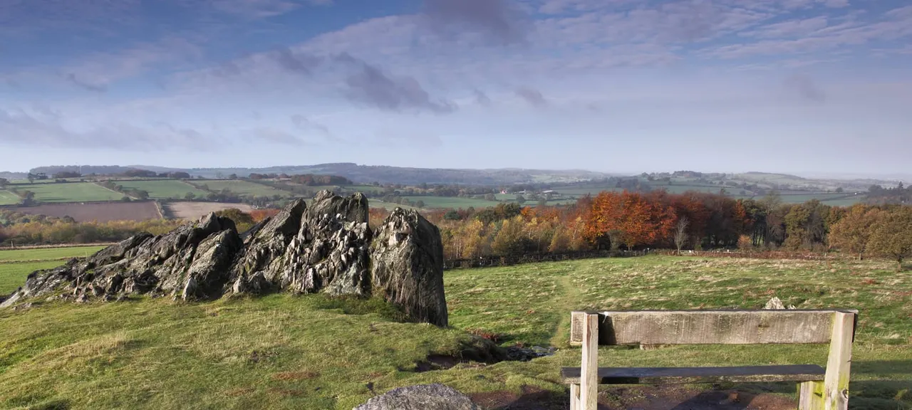 Wooden bench overlooking autumn countryside with distinctive rock outcrop, fields and wooded hills in distance.