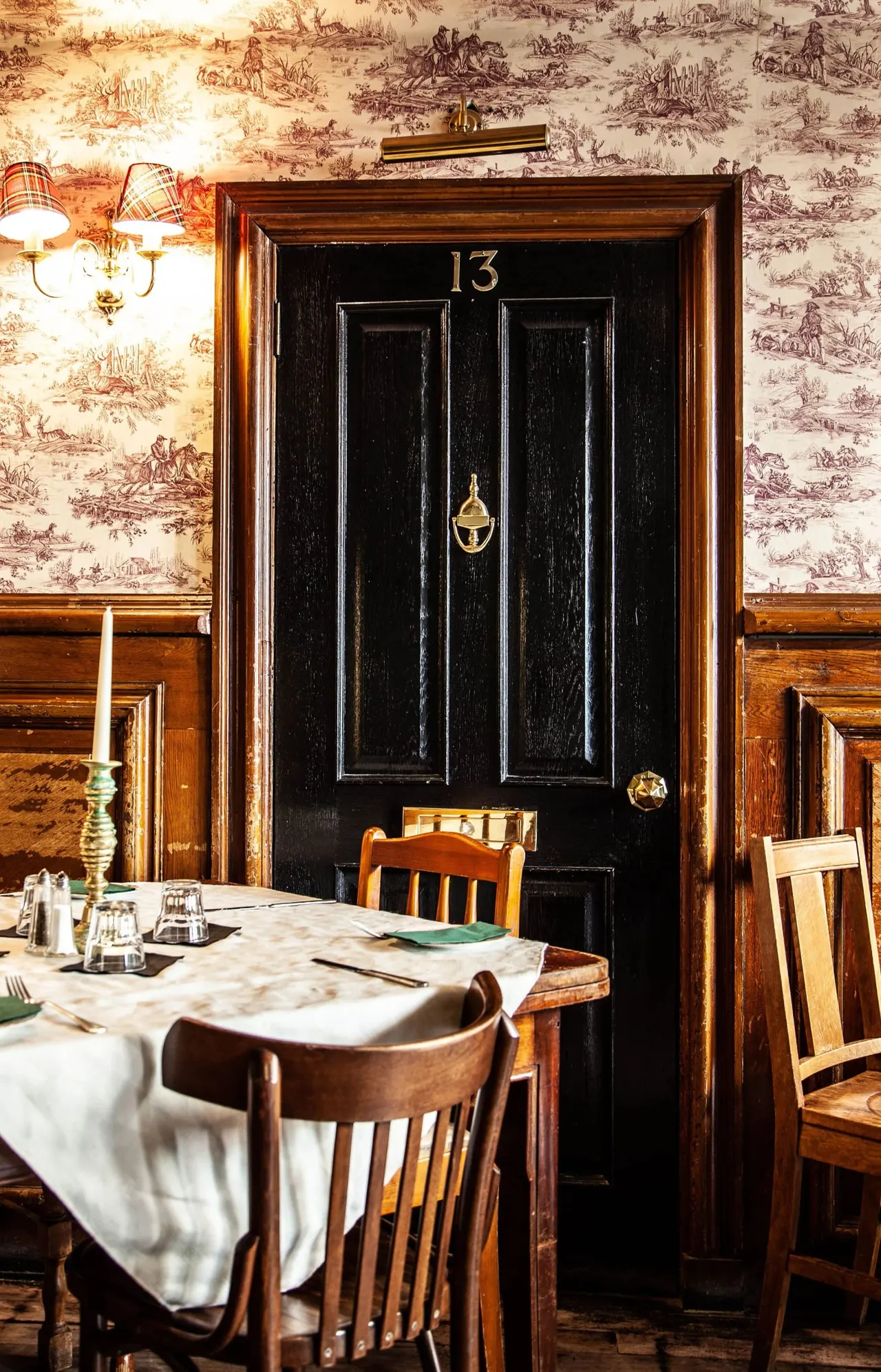 A dining room with wooden chairs at tables set with white linens, facing a black door numbered 13 with toile wallpaper and vintage lighting.