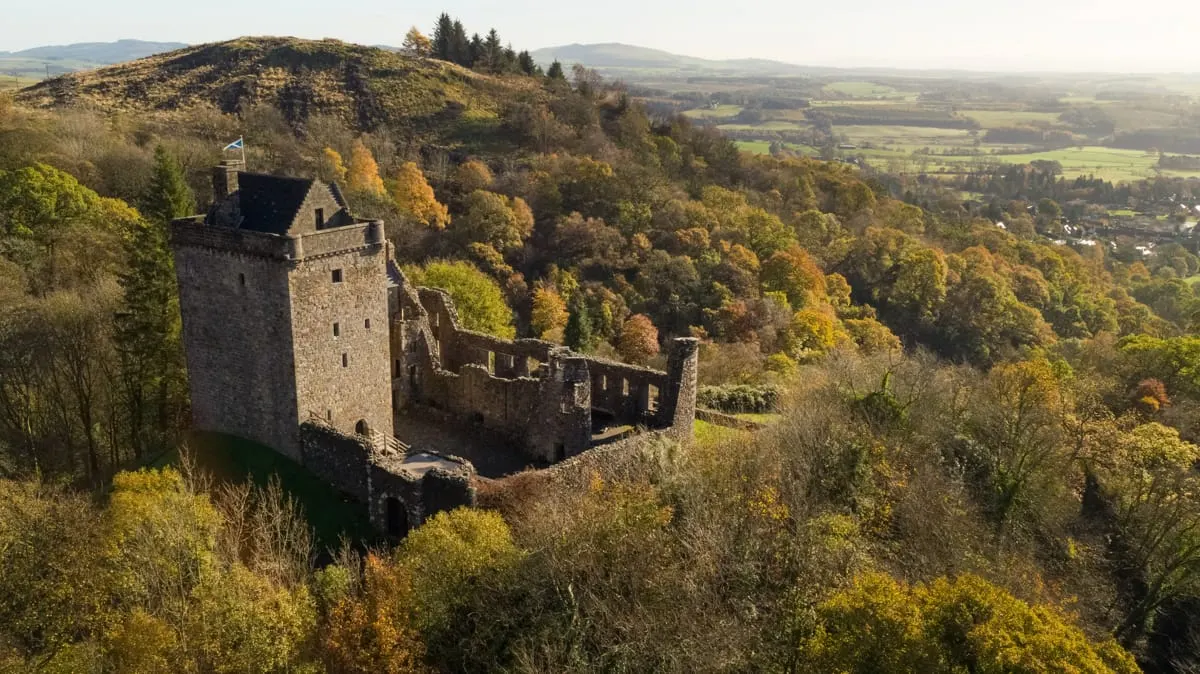 Ruined stone castle perched on a forested hilltop with expansive rural landscape and mountains visible beyond.