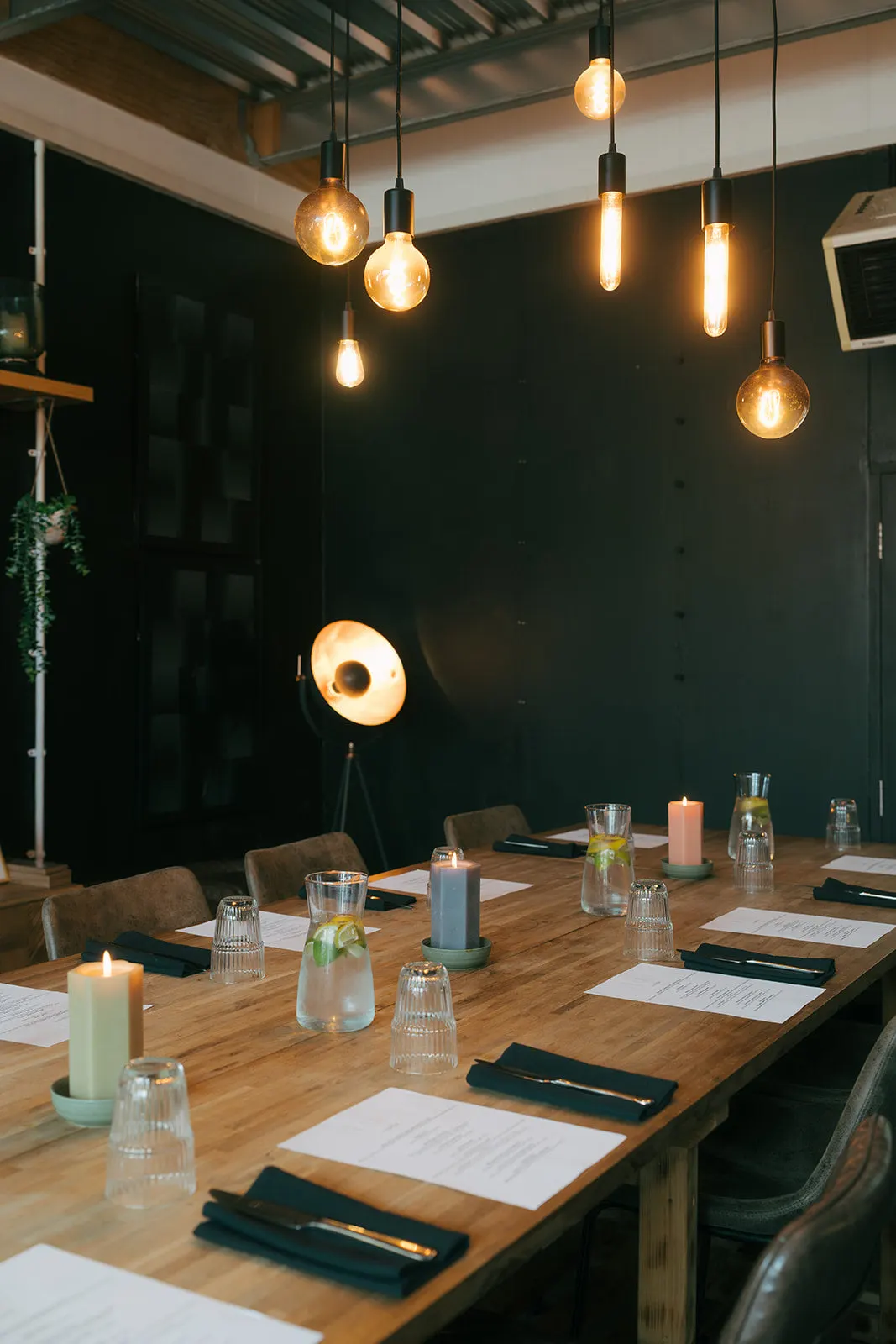 A dining room with a long wooden table set for a meal, with pendant lights overhead and dark walls.