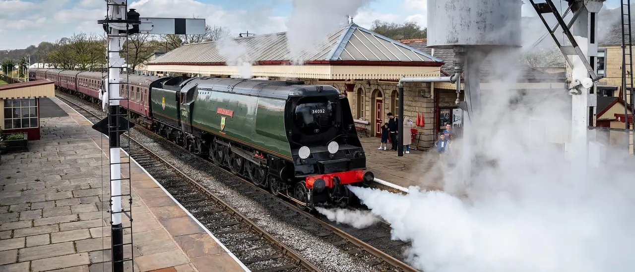 A heritage steam locomotive with maroon passenger carriages billows smoke whilst departing from a railway station platform, with onlookers watching nearby.