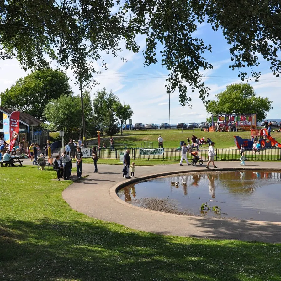 Families and visitors enjoying a sunny day at an outdoor leisure venue with a pond, grassy areas, and play facilities.