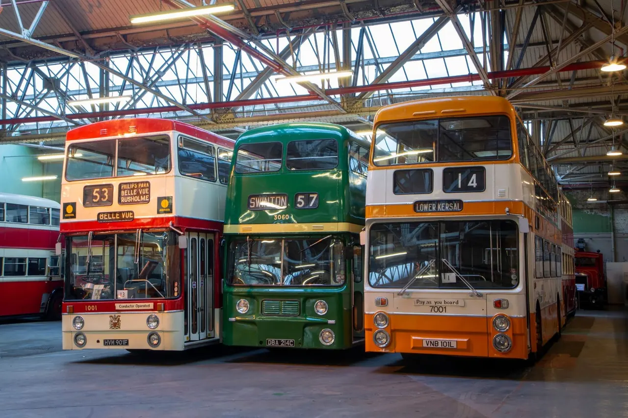 Three vintage double-decker buses in red, green and orange parked inside an industrial heritage hall with steel framework.