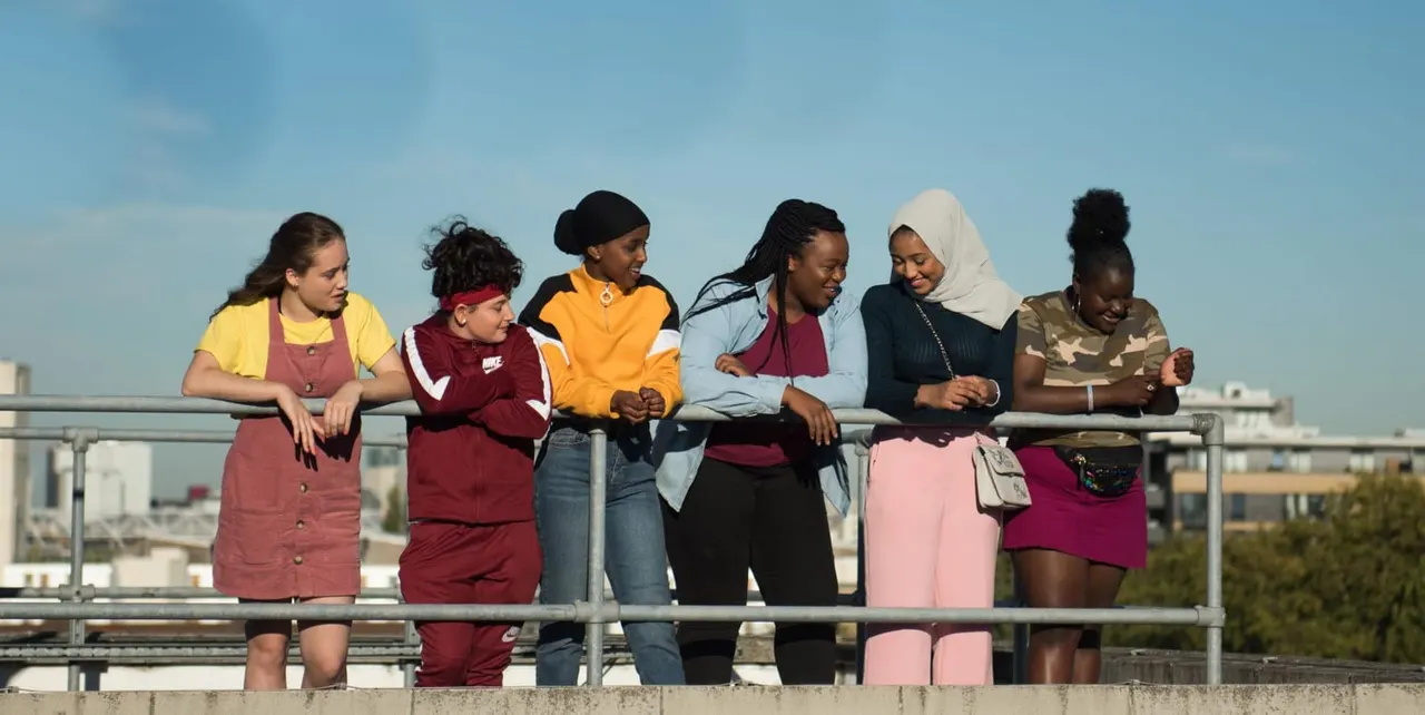 Six young people stand together on a rooftop terrace against a clear blue sky, smiling and chatting.