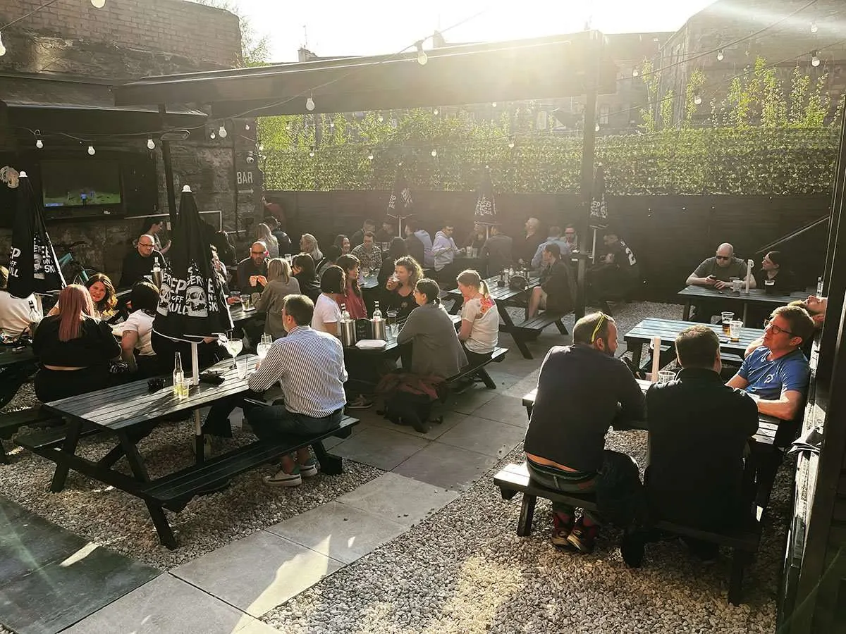 Busy outdoor beer garden with many patrons seated at picnic tables, drinking and socialising under a pergola with hanging lights.