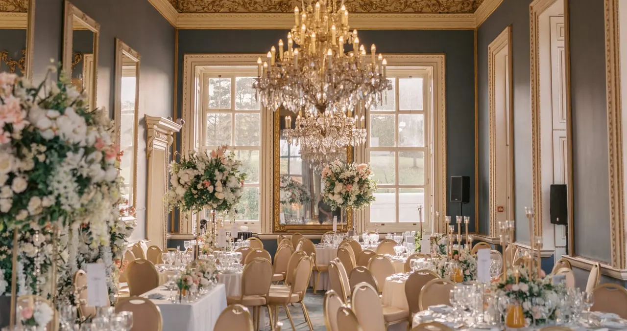Elegant ballroom set for a formal dinner with gold chairs, white tablecloths, floral centrepieces and a large crystal chandelier.