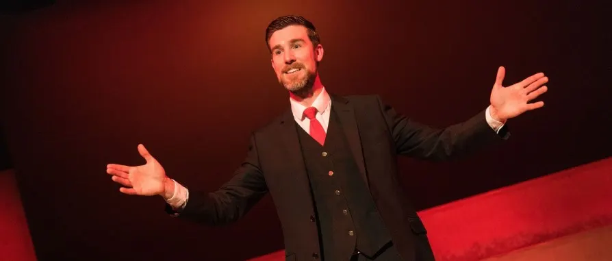 Man in formal black suit and red tie performing on stage with arms outstretched against red backdrop.