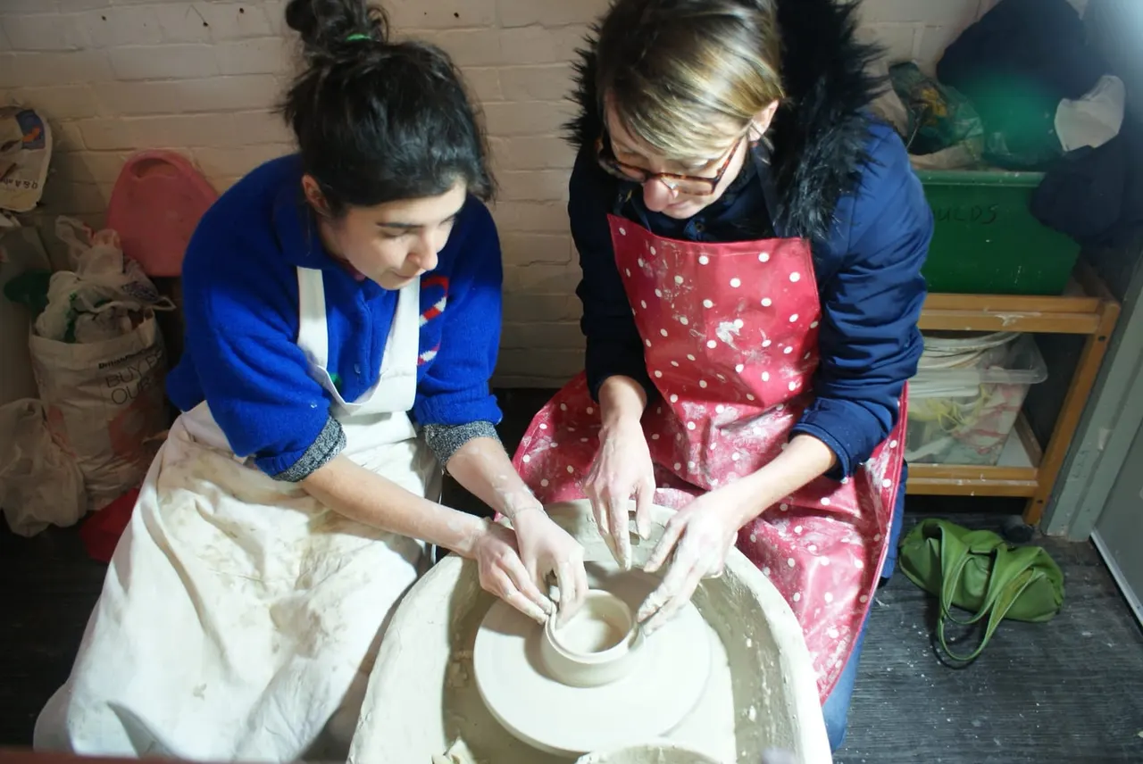 Two people working together at a pottery wheel, shaping clay during a hands-on pottery session.