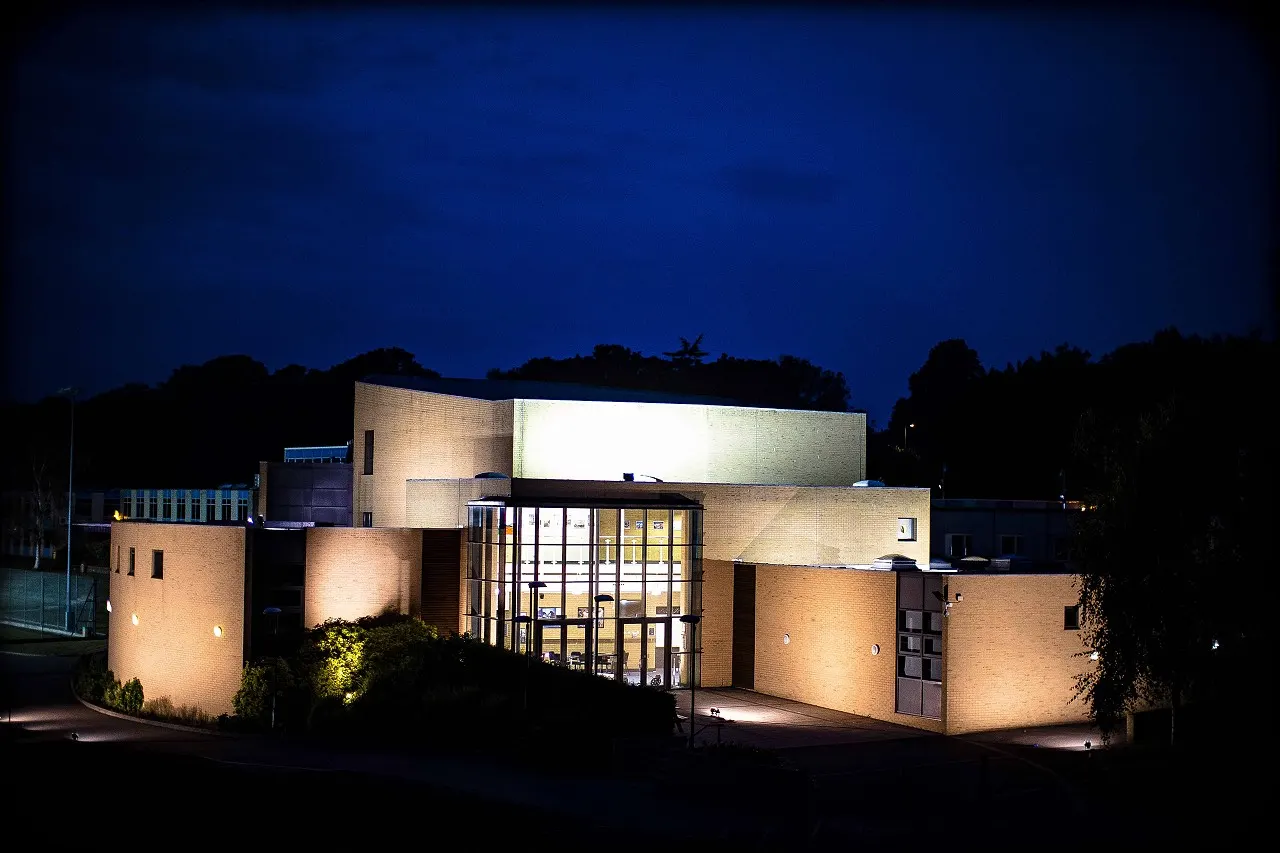Modern brick building with illuminated facade and large glass entrance doors at dusk, surrounded by trees.