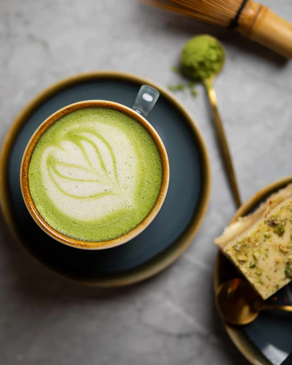 Matcha latte with latte art in a cup on a saucer, alongside matcha powder on a spoon and pastries.
