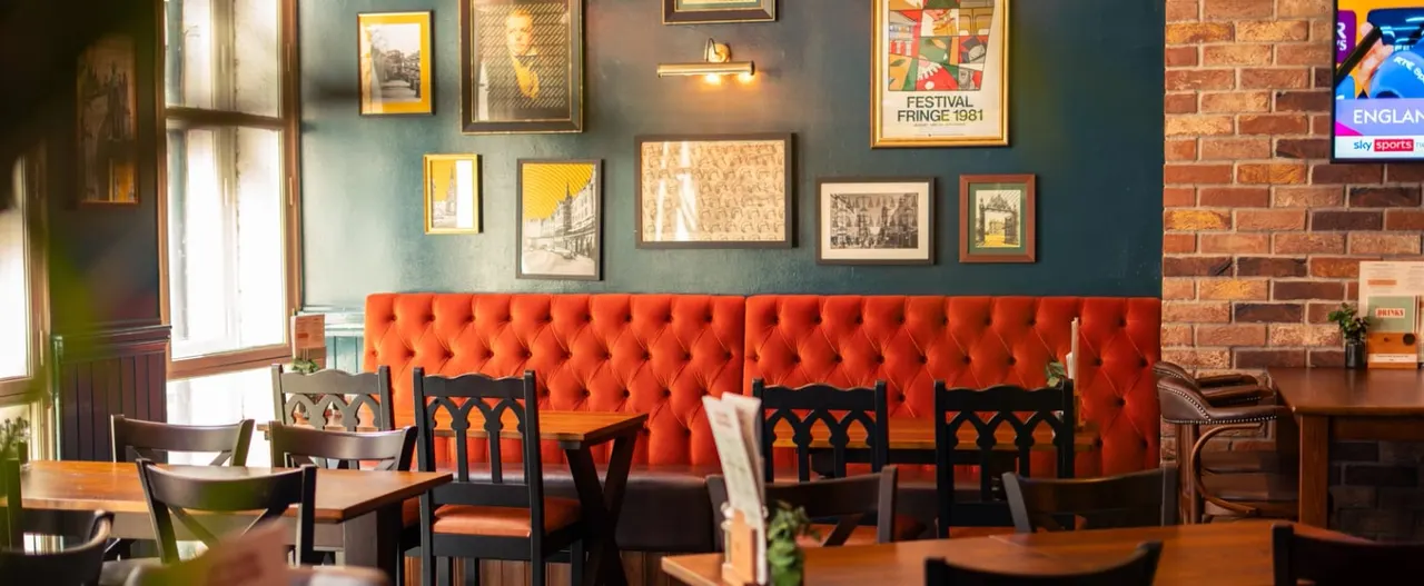 Interior of a pub with wooden tables, black chairs, a bright red quilted banquette, exposed brick wall and framed artwork.