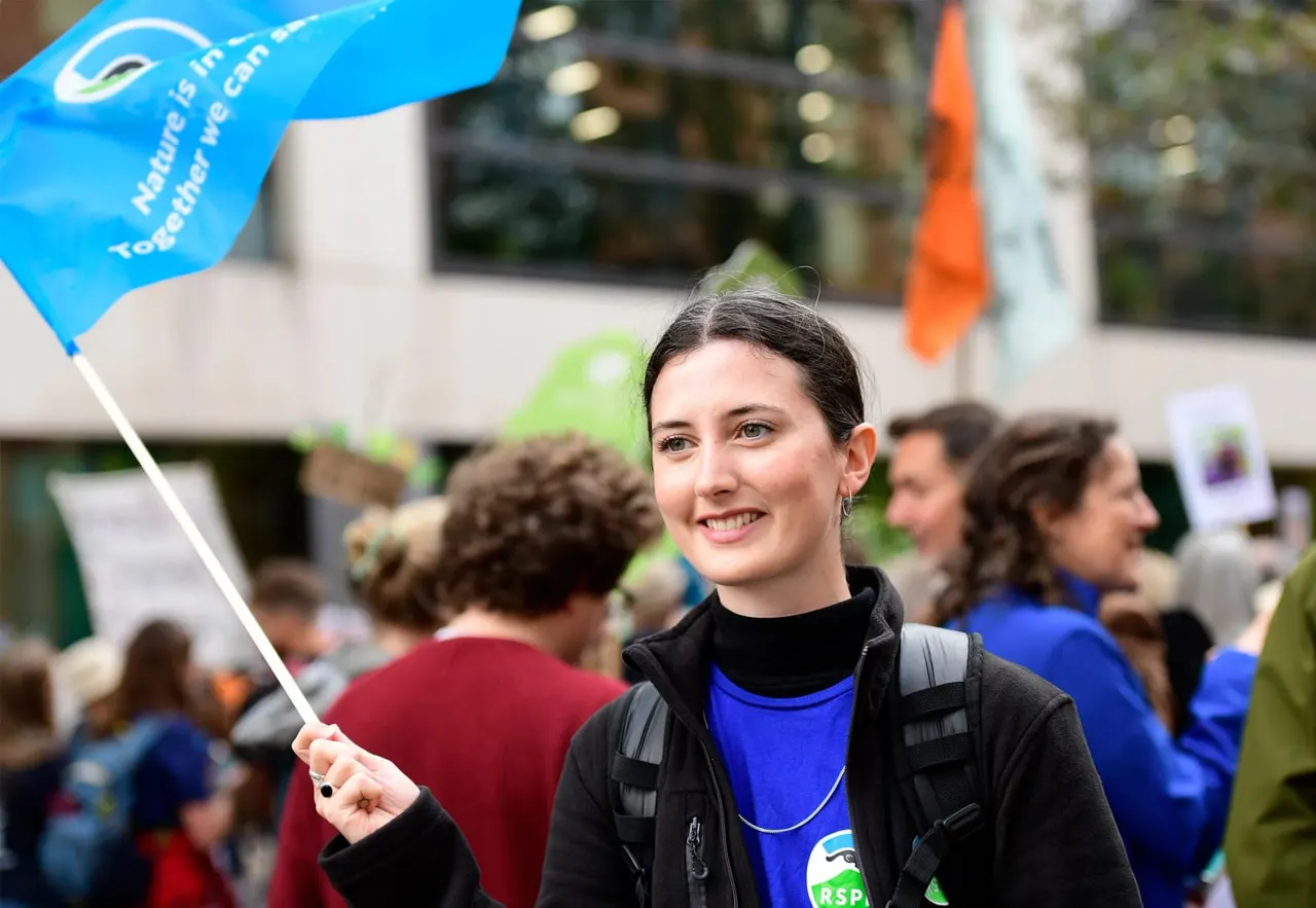 Woman holding blue flag at outdoor event with other people and visitors in background.