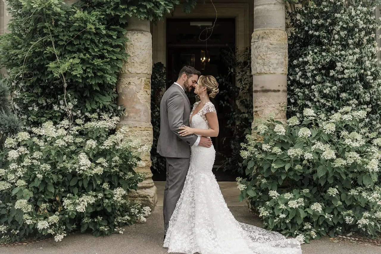 Bride and groom embrace in a stone archway framed by climbing white flowering plants at a garden venue.