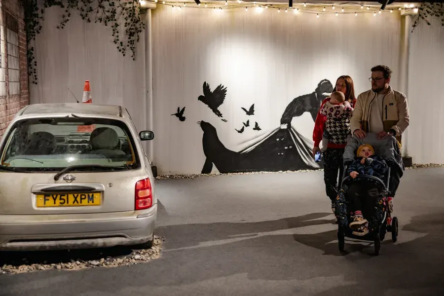 Family with pushchair posing beside a parked car in front of street art mural featuring silhouetted figures and birds.
