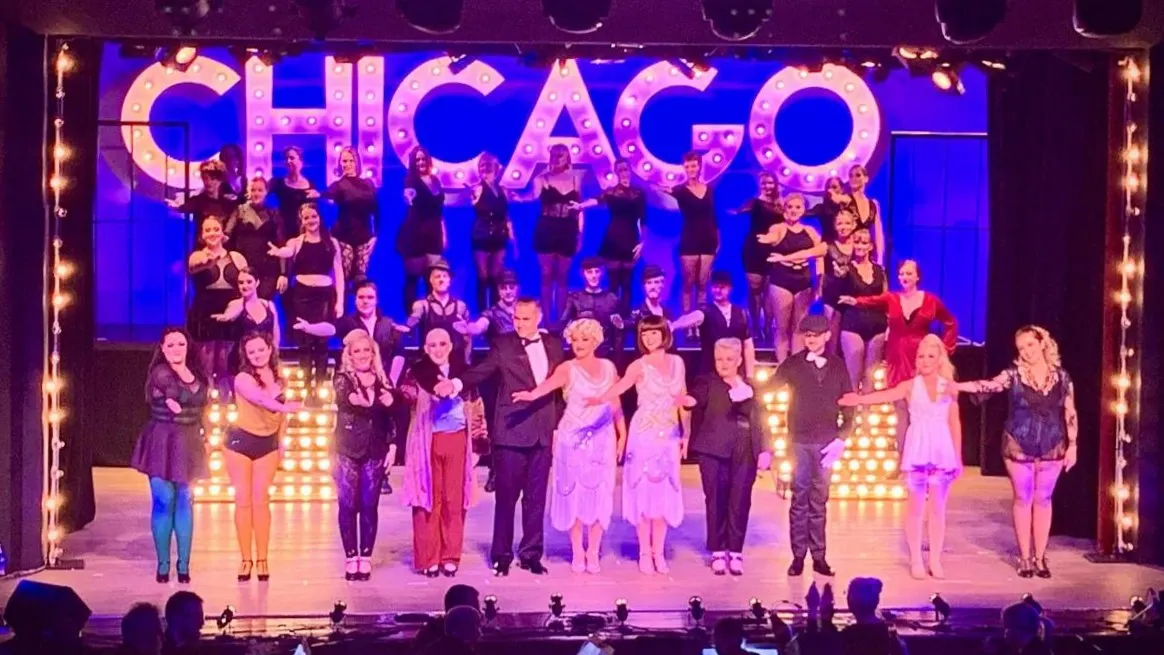 Large cast on stage during musical theatre production with illuminated "CHICAGO" sign and audience silhouettes in foreground.