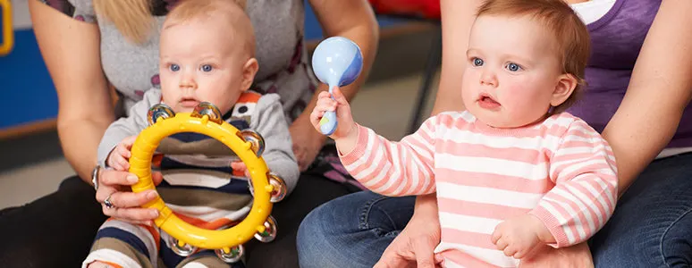 Two babies sit with adults, each holding colourful toy instruments—one with a yellow tambourine, one with a blue shaker.
