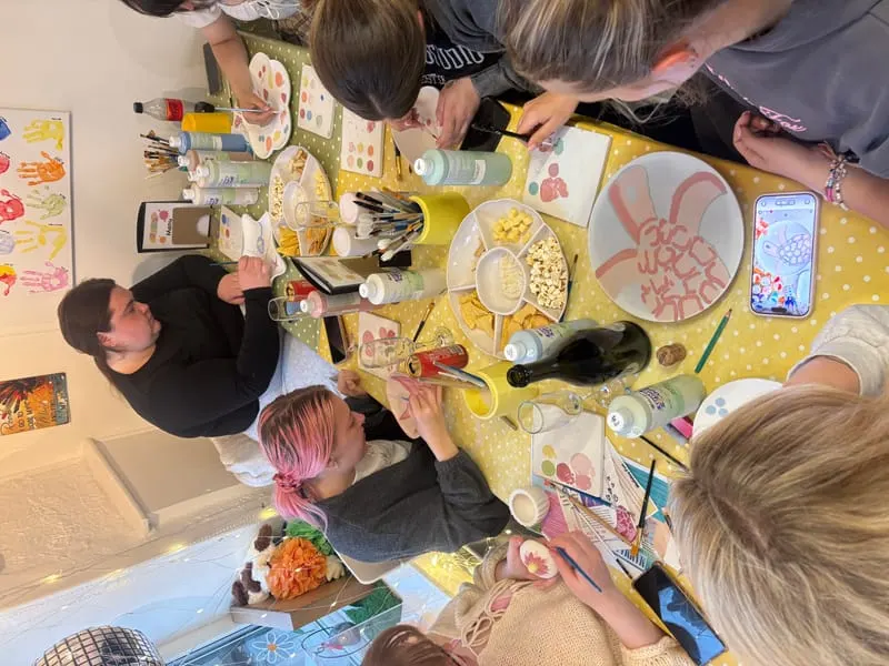 Children and adults gathered around a yellow table doing arts and crafts with paints, brushes, and ceramic pieces.