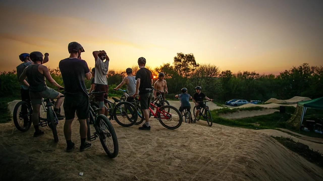 Group of cyclists with bikes gathered on a dirt track at sunset, with trees and earthen ramps in the background.