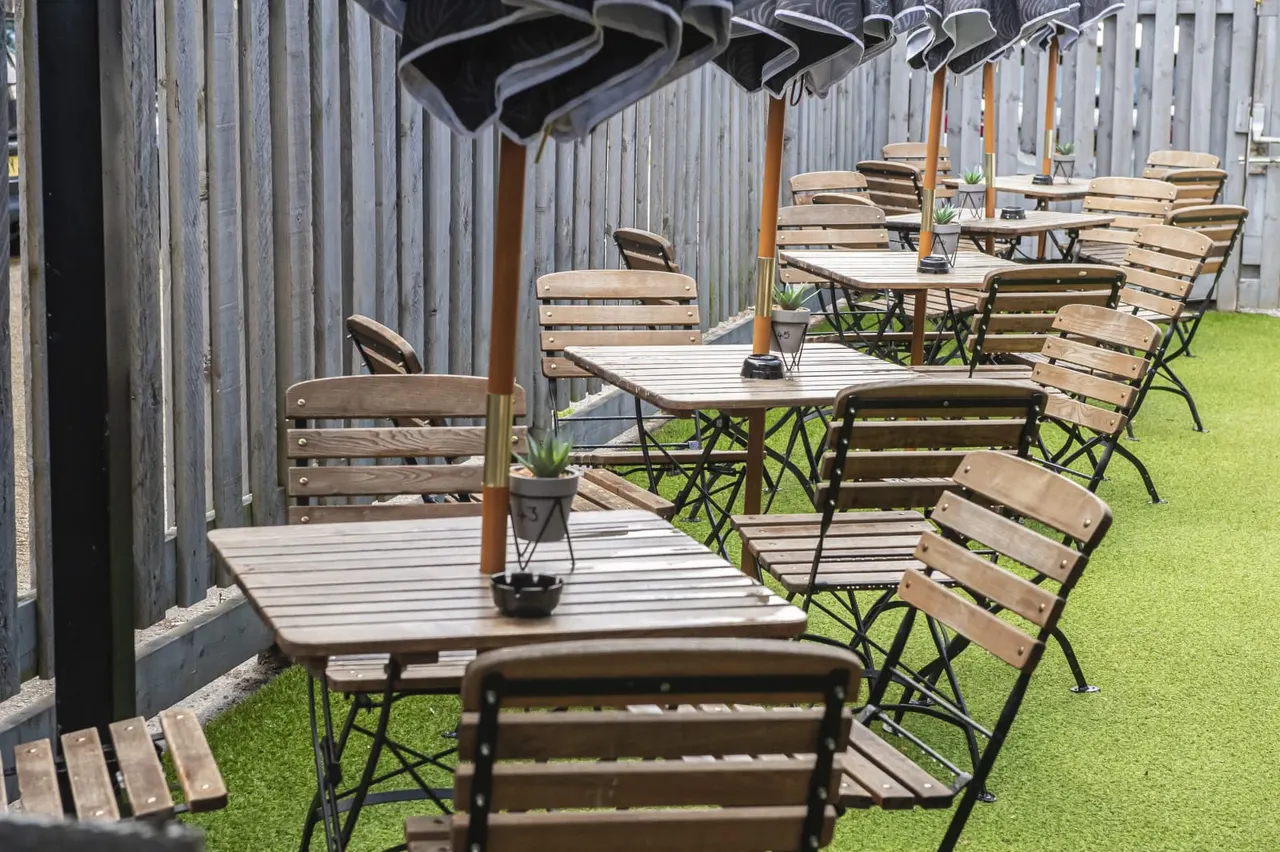 Outdoor garden seating area with wooden tables and chairs on artificial grass, shaded by umbrellas and wooden fencing.
