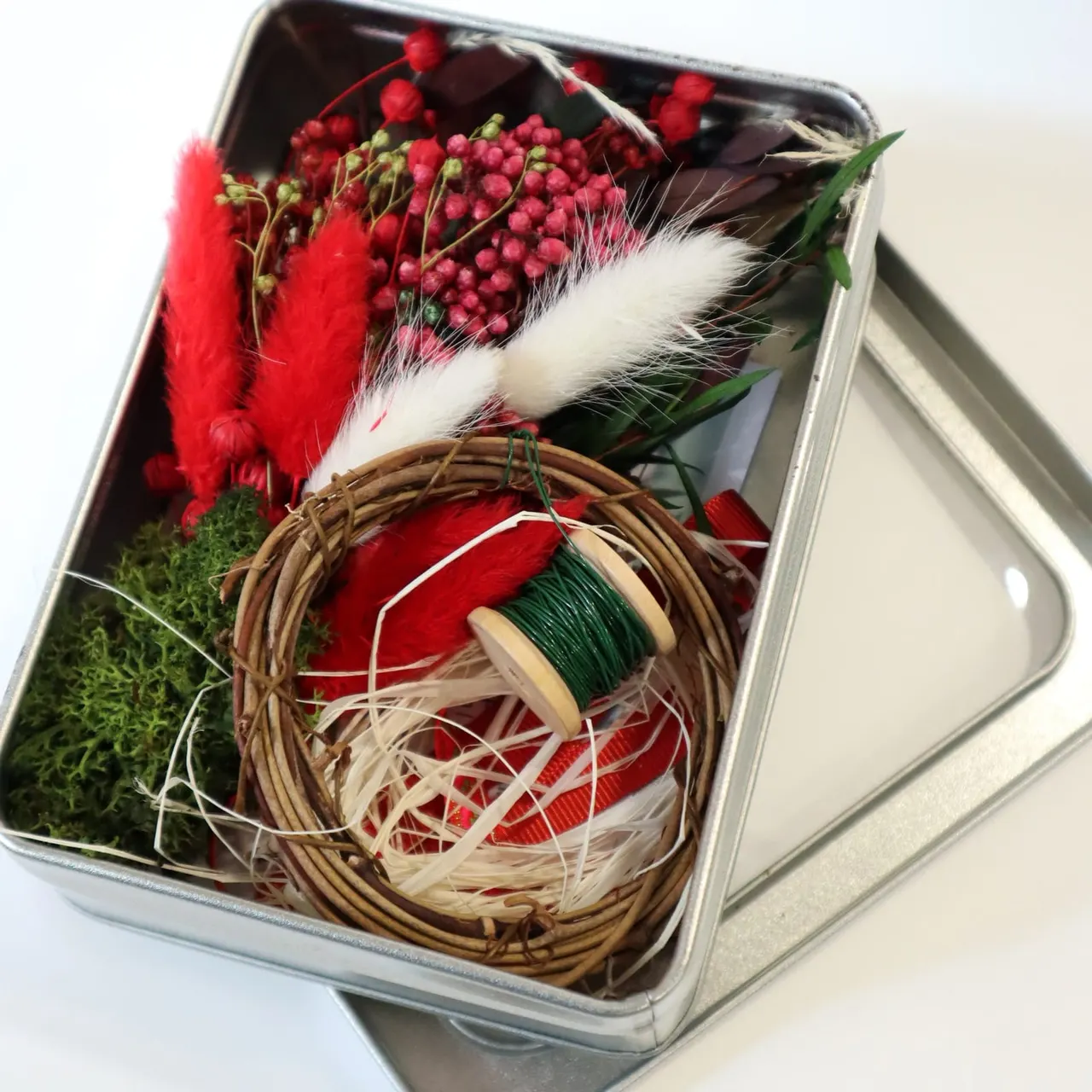 Decorative floral arrangement in a clear box with red dried flowers, red berries, white pampas grass, green foliage and a woven ring.