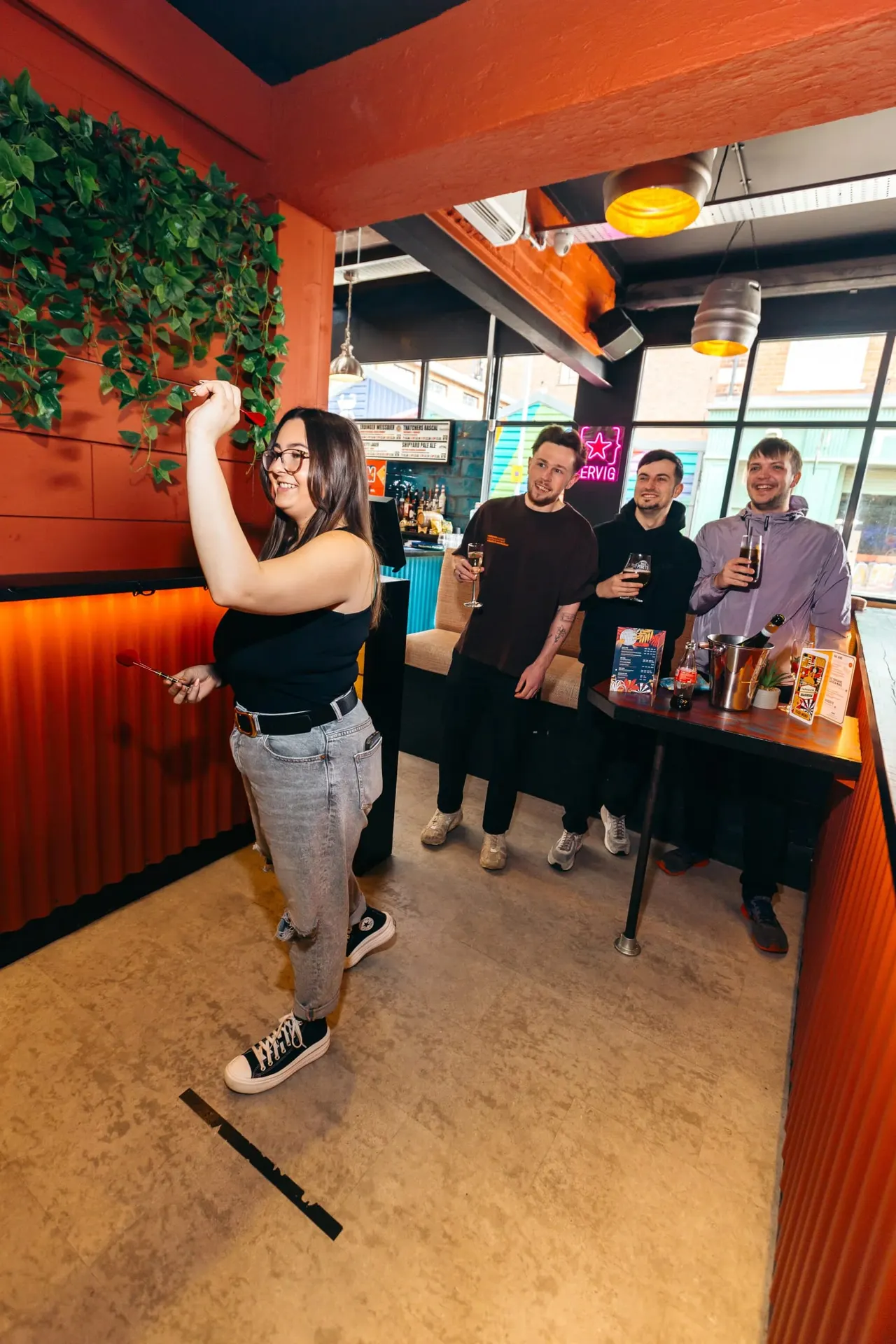 A woman plays darts whilst four men stand at a bar holding drinks in a modern venue with bright orange walls and hanging plants.