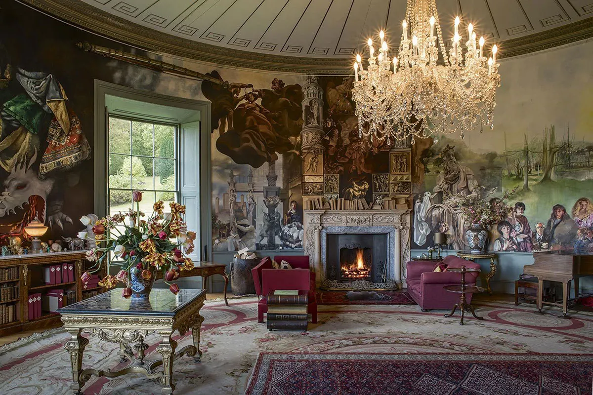 A grand oval room with ornate ceiling, crystal chandelier, decorative murals, fireplace and burgundy upholstered furniture, empty of people.