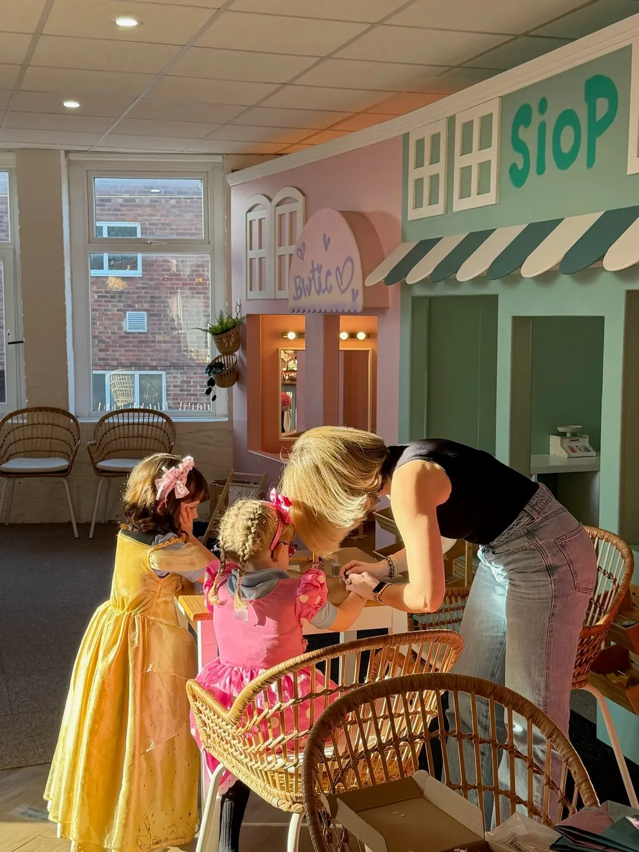 Adult and two young children playing at a shop-themed play area with colourful painted storefronts and props.