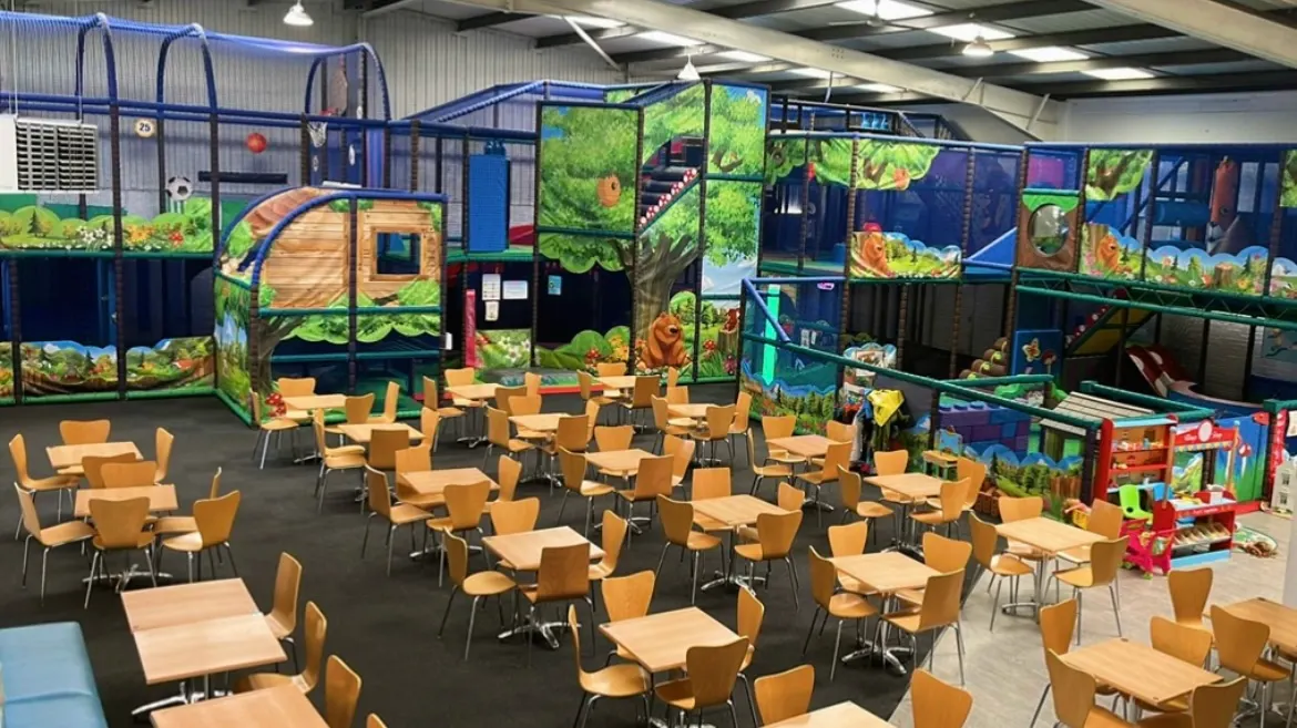 Indoor play centre with wooden chairs and tables in foreground, large colourful play structures with slides and climbing frames decorated with nature murals in background.