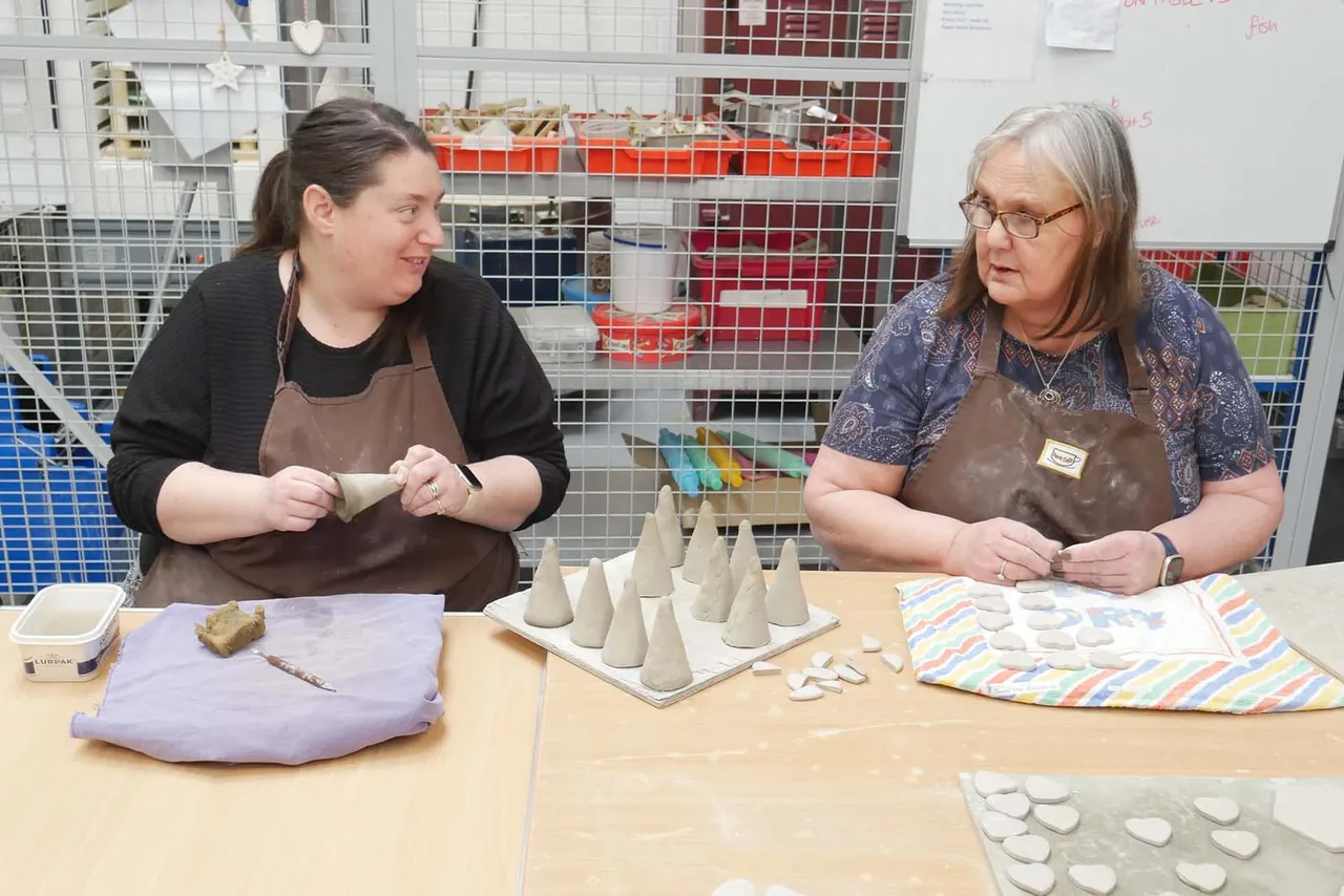 Two women work together at a pottery table, hand-building clay vessels with shelving of ceramic pieces visible behind them.