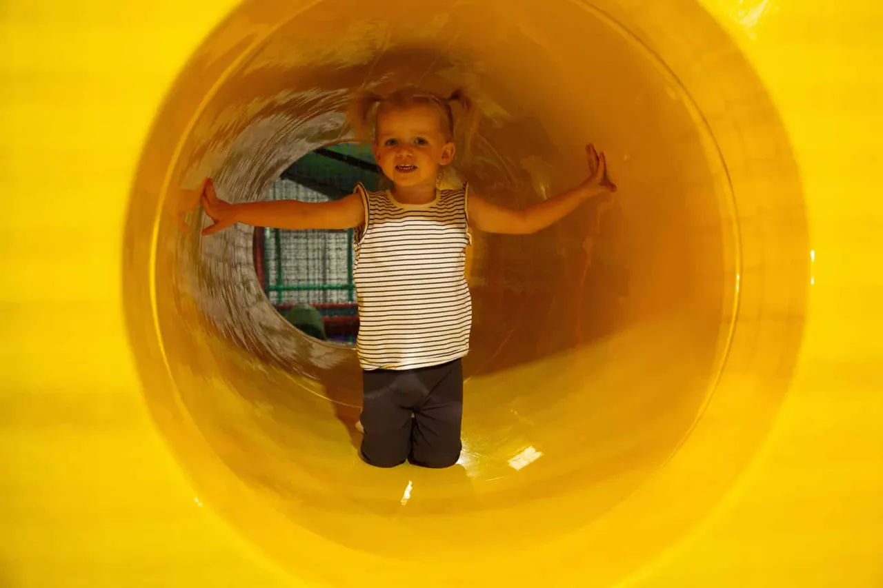 A young child stands inside a bright yellow play tunnel with arms outstretched, smiling.