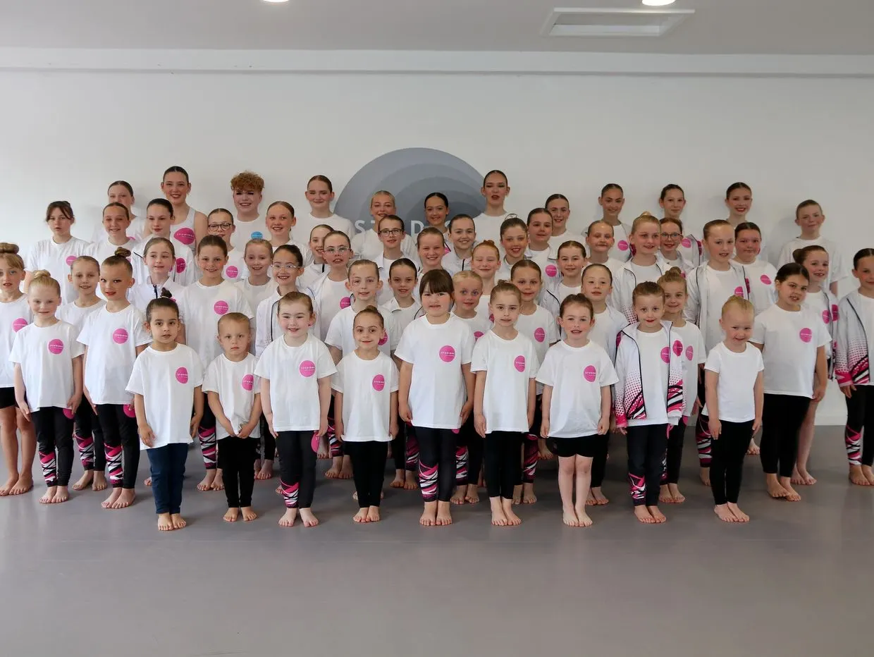 Large group of children in white t-shirts and patterned leggings standing barefoot in a dance studio.