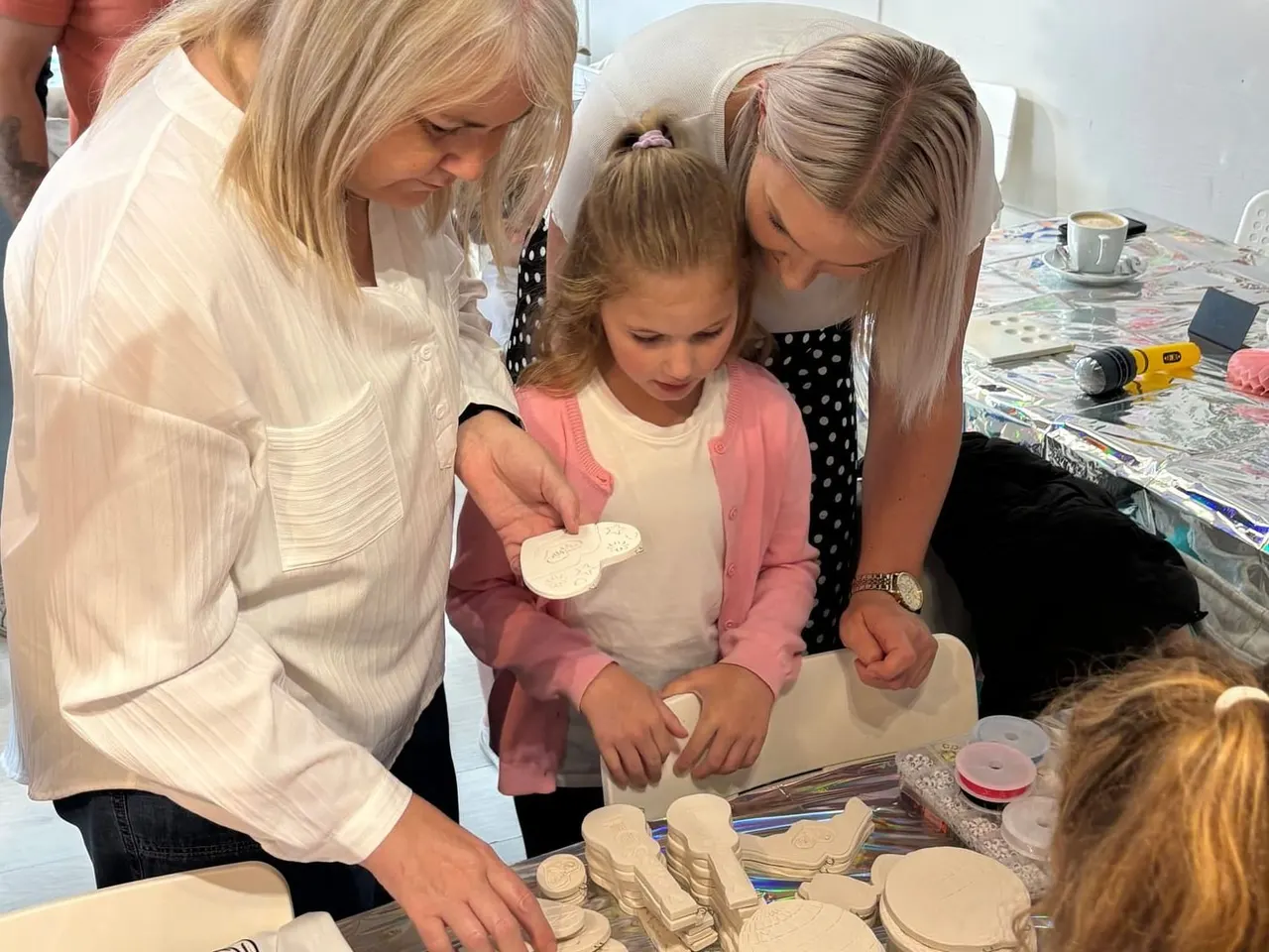 Two women and a young child painting ceramic items at a craft table during a pottery painting session.
