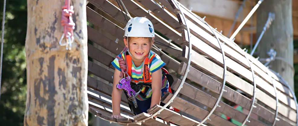 Child wearing safety helmet and harness navigating wooden climbing structure outdoors.