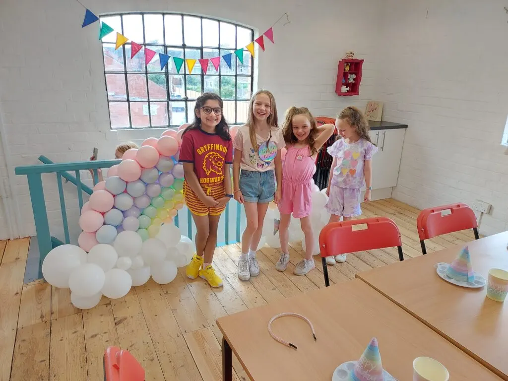 Five children posing together at a decorated party venue with a colourful balloon arch and bunting.