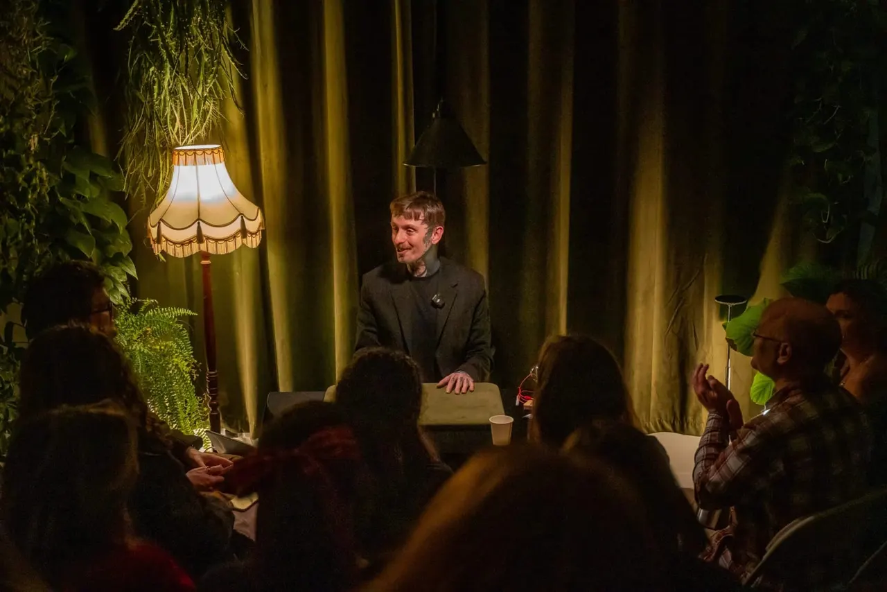A man performs a magic trick on stage surrounded by an audience in an intimate theatre setting with decorative lighting and vintage furnishings.