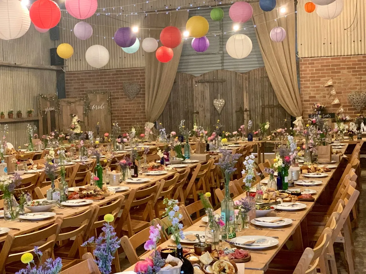 Decorated barn venue with long wooden tables set for a wedding reception, featuring colourful paper lanterns overhead, floral centrepieces, and place settings ready for guests.