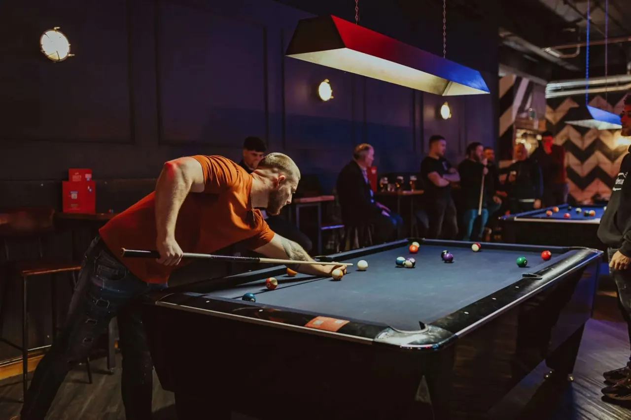 Man in orange shirt playing pool at a table whilst others socialise in a dimly lit entertainment venue.