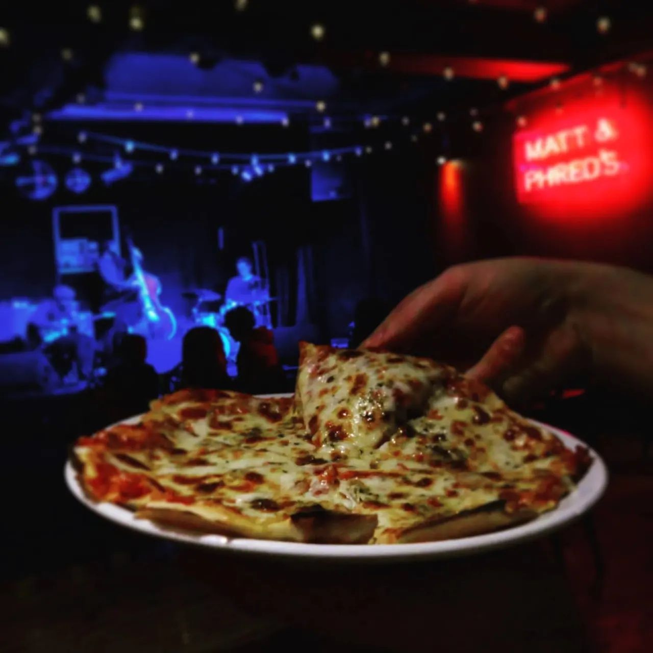 A hand holds a slice of pizza in front of a plate at a lively jazz club with blue-lit stage and red neon signage.