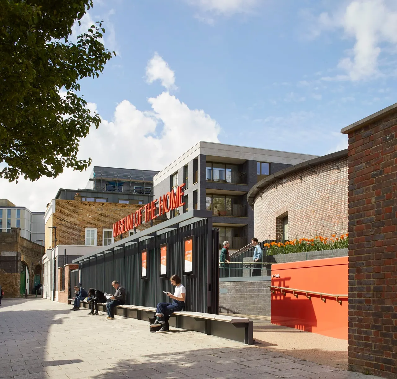Visitors sit on benches outside a modern museum building with red signage and brick architecture on a sunny day.