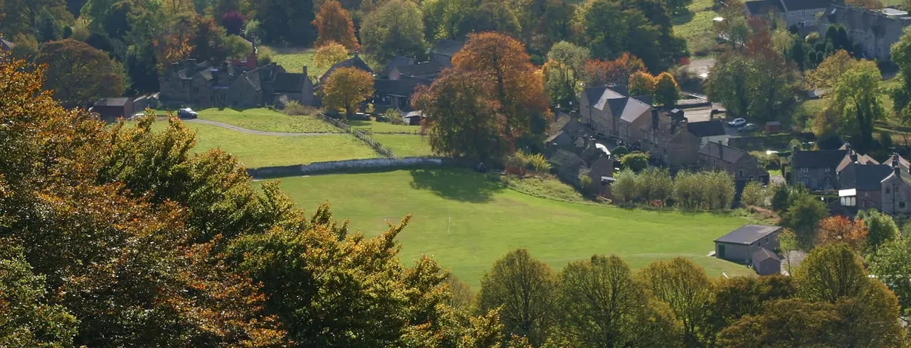 Aerial view of a rural valley village with stone buildings, green fields, and trees in autumn colours.