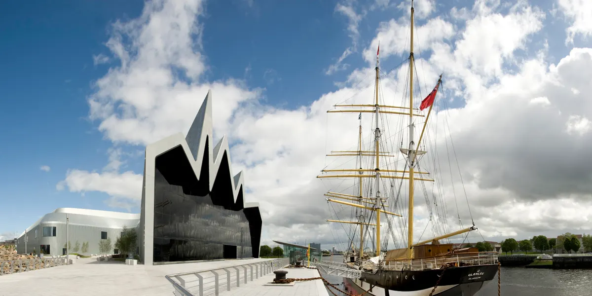 Historic sailing ship moored beside modern riverside museum building under blue sky with white clouds.